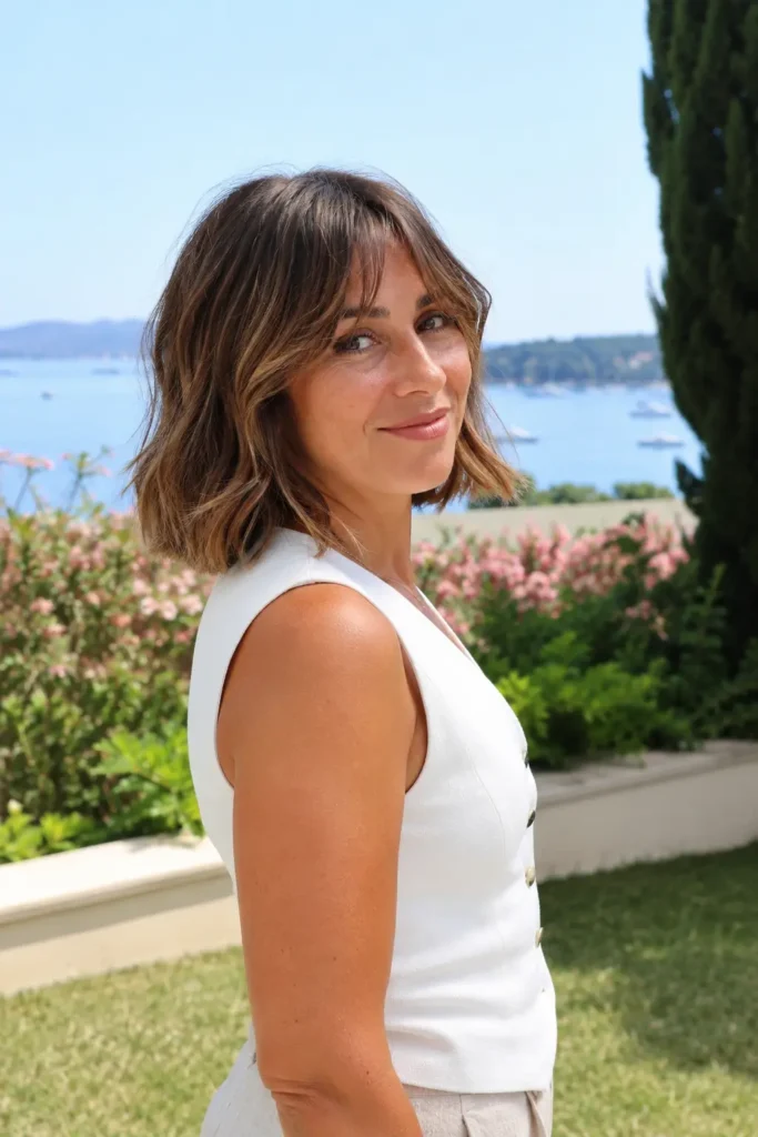 woman aged 25–45 with a beachy bob hairstyle and soft curtain fringe, rich brown hair with subtle highlights, standing in a seaside garden. She wears a casual elegant white outfit and looks over her shoulder toward the camera