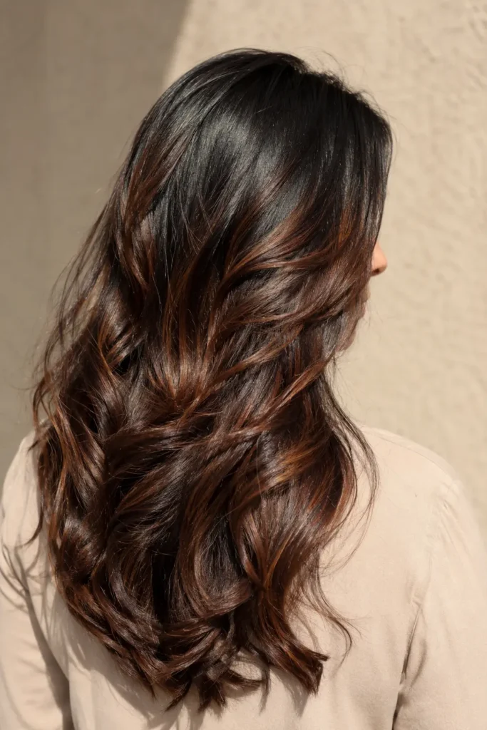 woman aged 30–50 with long black hair enhanced by deep maple brown highlights, shown mostly from the back with a gentle side profile and minimal facial visibility. Her glossy,