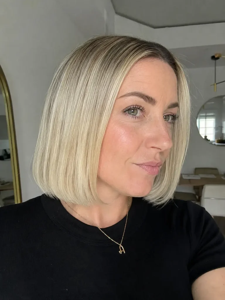 Close-up portrait of a real English woman with a sleek blunt chin-length bob haircut in glossy light blonde hair. The hairstyle is the focal point, showcasing smooth, straight strands with a polished finish and precise cut line at the jaw