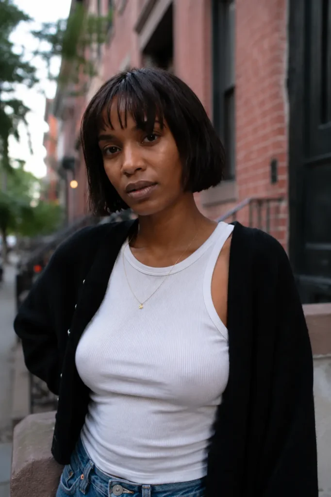 Black woman with a sleek dark brunette bob haircut and choppy bangs standing outdoors in an urban setting. Her hairstyle is neatly styled and clearly visible, with natural-looking hair texture and realistic skin detail. She wears a stylish casual outfit and a subtle necklace