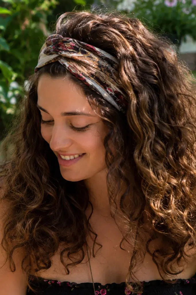 outdoor portrait of a woman wearing a boho-style fabric headband with long, soft natural curls. Her authentic facial features and natural hair texture are highlighted,