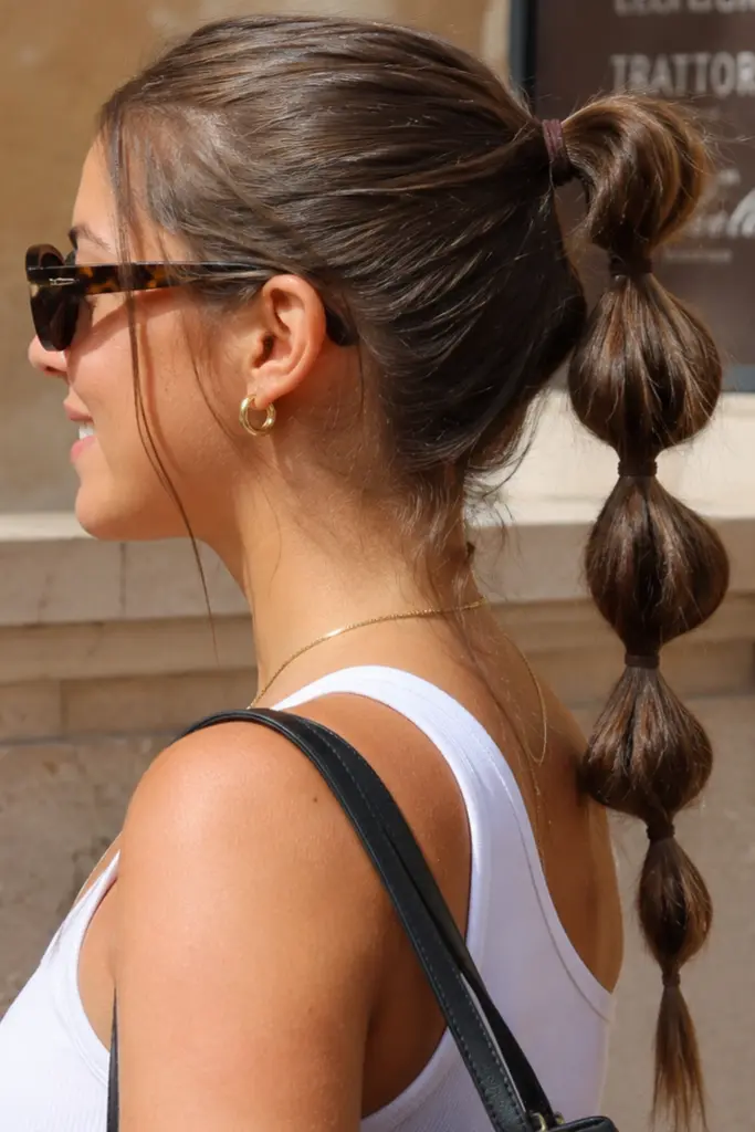 Side-profile portrait of a woman with natural brunette hair styled in a bubble ponytail, wearing sunglasses and a white tank top in bright summer sunlight