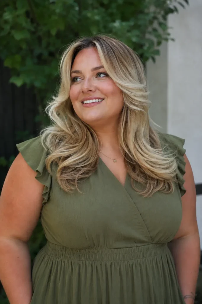 Plus-size English woman with a butterfly haircut and curtain bangs, featuring dimensional blonde balayage and voluminous layered hair. She wears a modest olive-green dress with a covered neckline,
