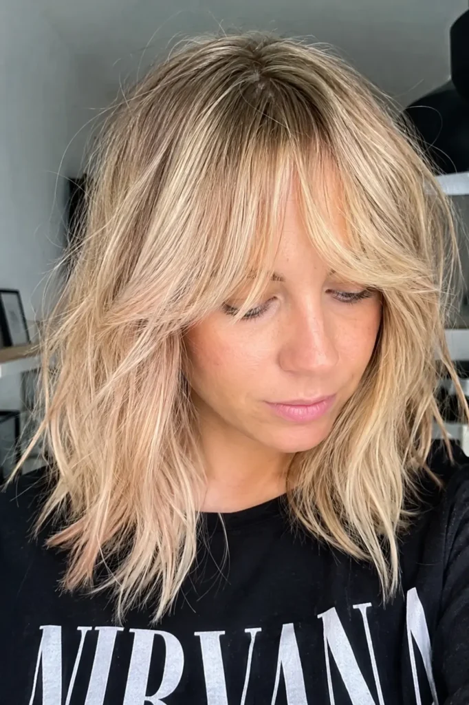 English woman in her late 20s to 30s with a choppy layered haircut and long curtain fringe, wearing her warm champagne blonde hair in a tousled shoulder-length style