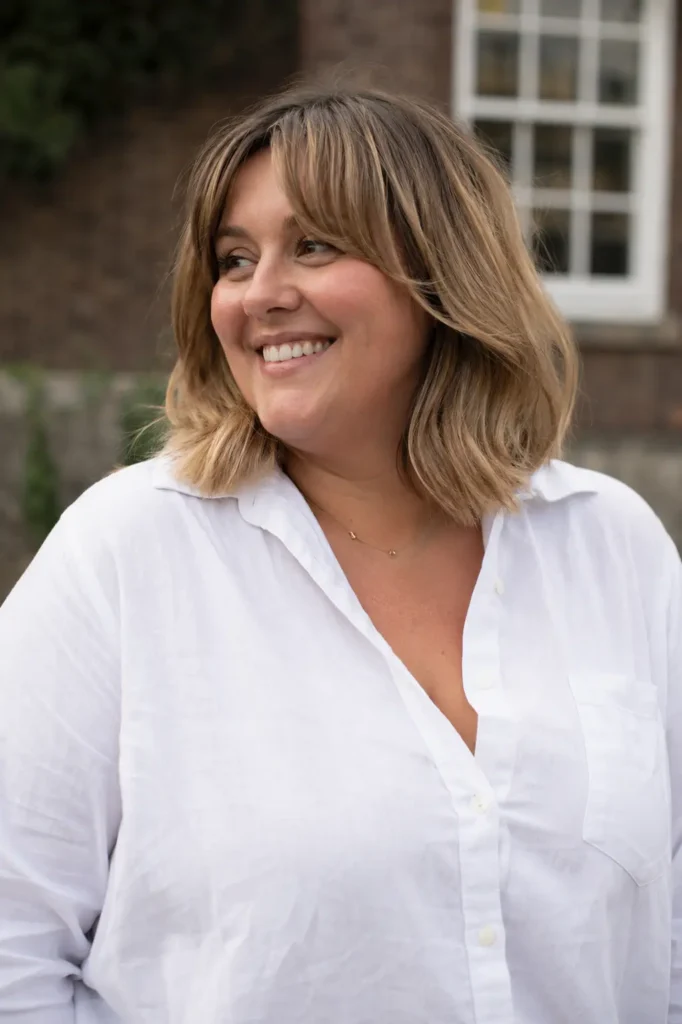 Plus-size English woman with a collarbone-length haircut featuring curtain bangs and soft blonde balayage highlights, wearing a modest white button-up shirt