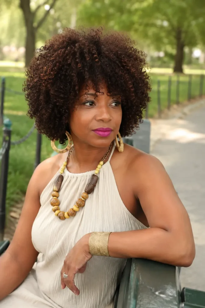 Black woman aged 30–55 with a shaped curly afro hairstyle featuring voluminous, defined natural curls. She is seated in a sunlit outdoor park wearing an elegant sleeveless cream outfit and statement jewelry