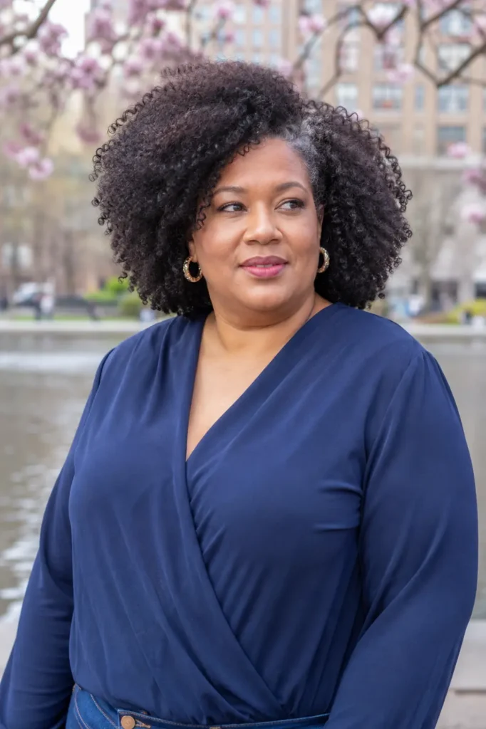 plus-size woman aged 30–60 with a curly afro hairstyle and deep side part, showcasing voluminous natural curls with visible texture and definition. She wears an elegant navy wrap-style top and stands in an outdoor city park near a waterfront with blooming trees