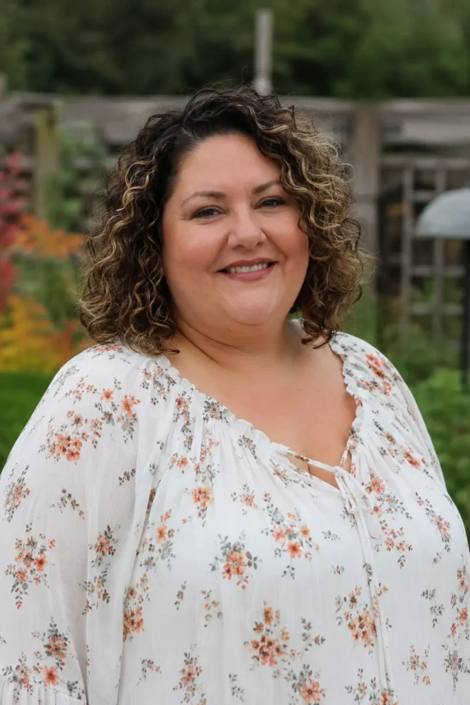 Close-up portrait of a real plus-size woman aged 30–55 with a curly bob hairstyle and face-framing blonde highlights. Her defined natural curls frame her face beautifully, with the hairstyle prominently displayed. She wears an elegant floral blouse