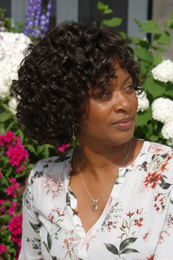 Black woman aged 30–50 with a layered curly bob hairstyle featuring voluminous, well-defined curls. She wears an elegant floral blouse and natural makeup, standing in a colorful outdoor garden filled with blooming flowers.