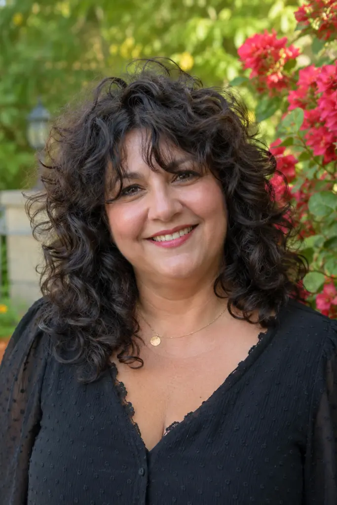 plus-size woman aged around 30–55 with a curly butterfly cut hairstyle featuring layered, voluminous curls. She wears an elegant black blouse and smiles warmly in a vibrant outdoor floral garden
