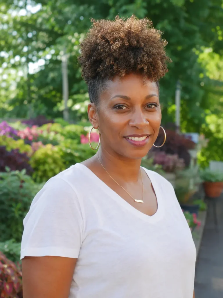 Black woman aged 25–50 with a curly crown cut hairstyle featuring elevated natural curls on top and closely tapered sides. She is wearing an elegant casual light-colored top and standing in a lush outdoor garden
