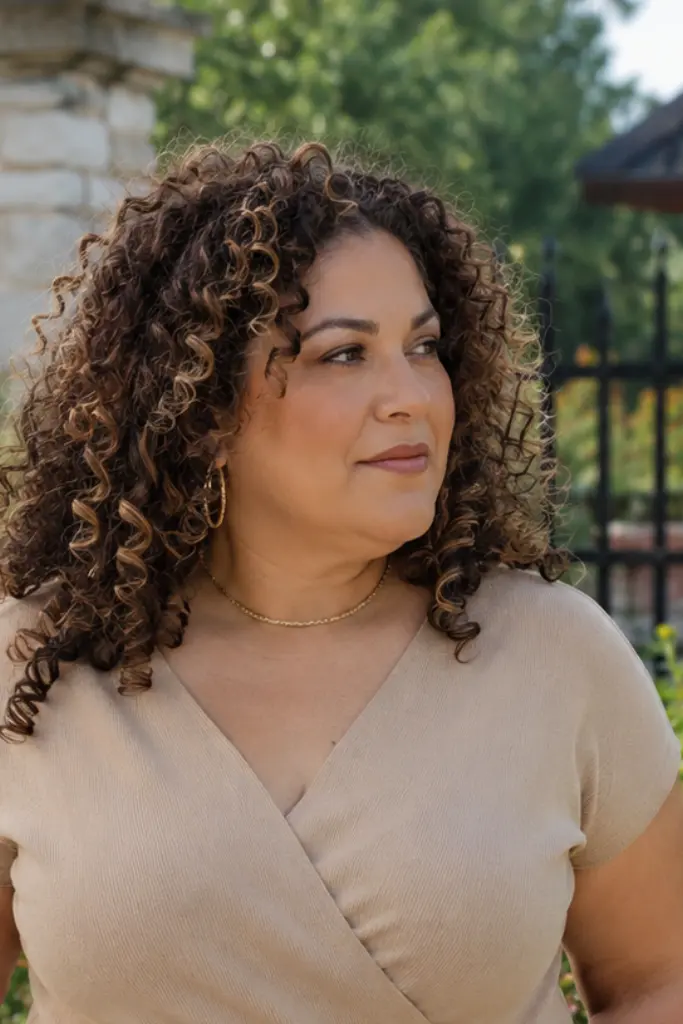 plus-size woman aged 30–55 with voluminous curly hair featuring warm caramel highlights, standing in a colorful outdoor garden. Her defined natural curls frame her face and are clearly visible, with realistic skin texture and an authentic appearance