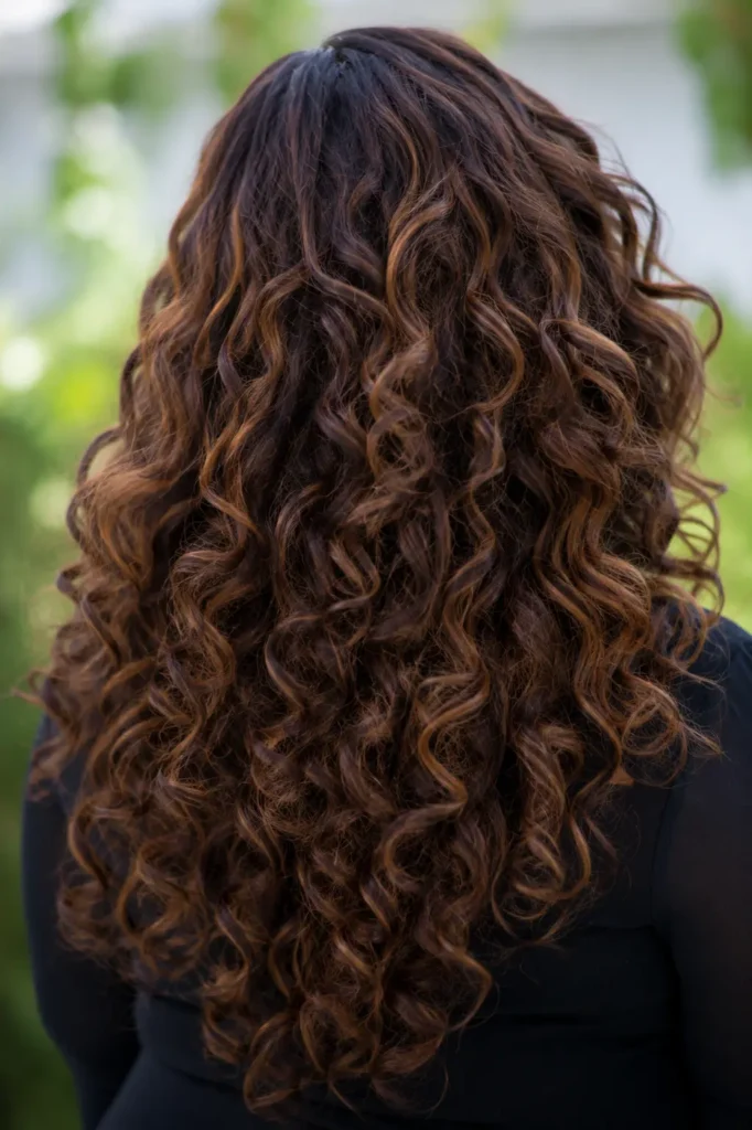 Close-up back view of a real plus-size woman with long curly hair and side layers, featuring cascading, well-defined natural curls with subtle caramel highlights
