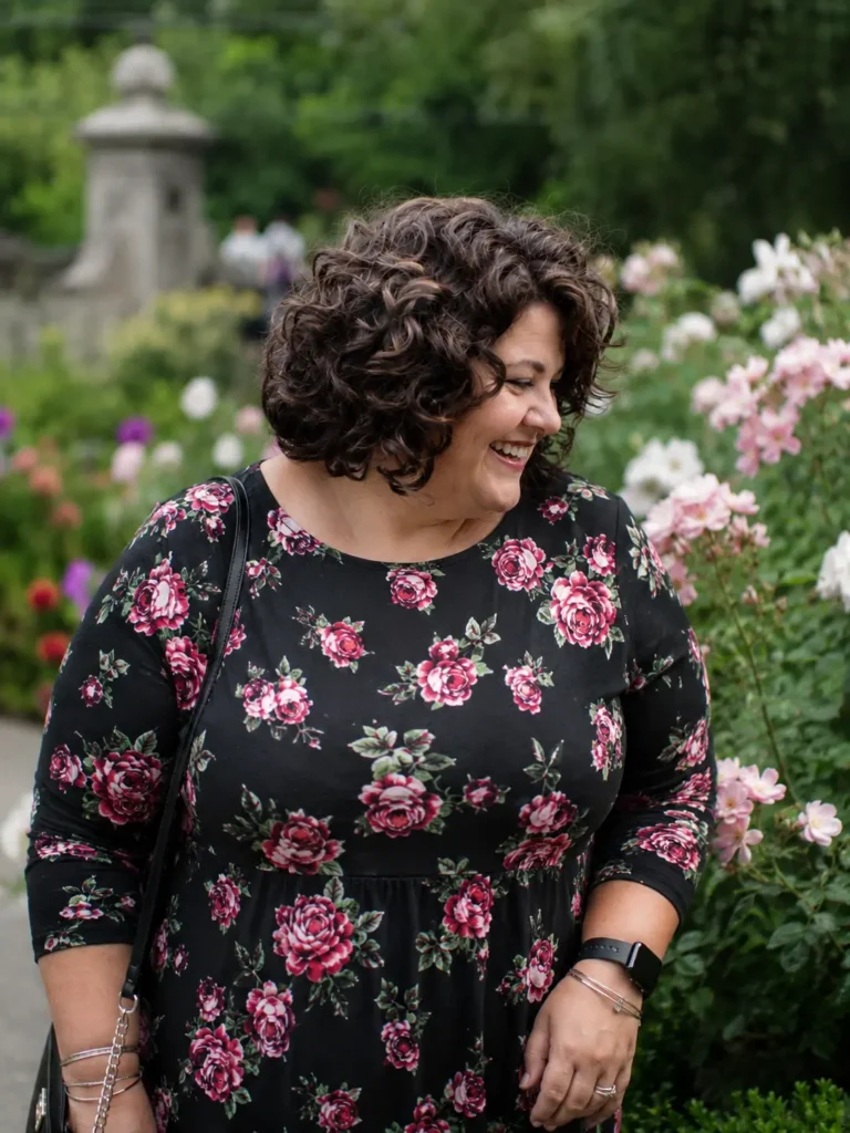 plus-size woman aged 30–55 with a layered curly bob hairstyle, featuring voluminous, well-defined curls and natural texture. She is wearing a modest floral dress with full chest coverage and standing in a blooming outdoor garden filled with pink flowers.
