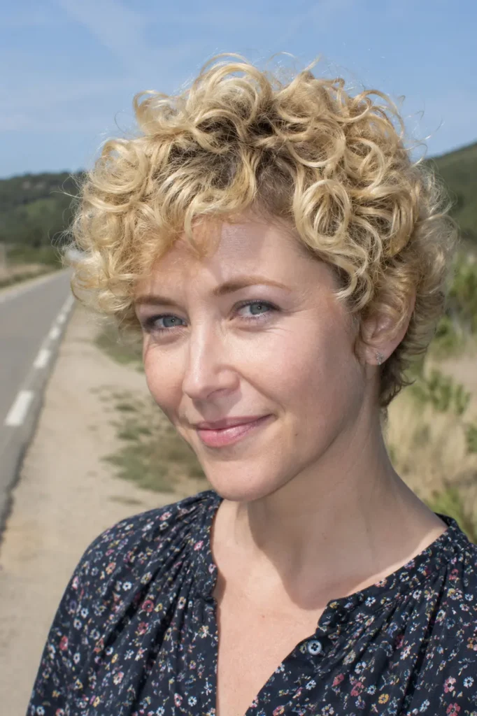 English woman with a curly pixie haircut featuring soft blonde curls and defined texture. The hairstyle is the main focus, with natural volume and detailed curl definition highlighted by bright daylight.