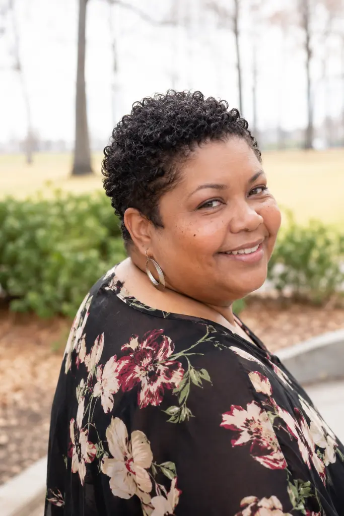 Close-up vertical portrait of a real plus-size woman aged around 30–60 with a curly TWA (teeny weeny afro) hairstyle featuring soft, natural coils. She is smiling warmly in an outdoor park setting with a softly blurred background of trees and greenery