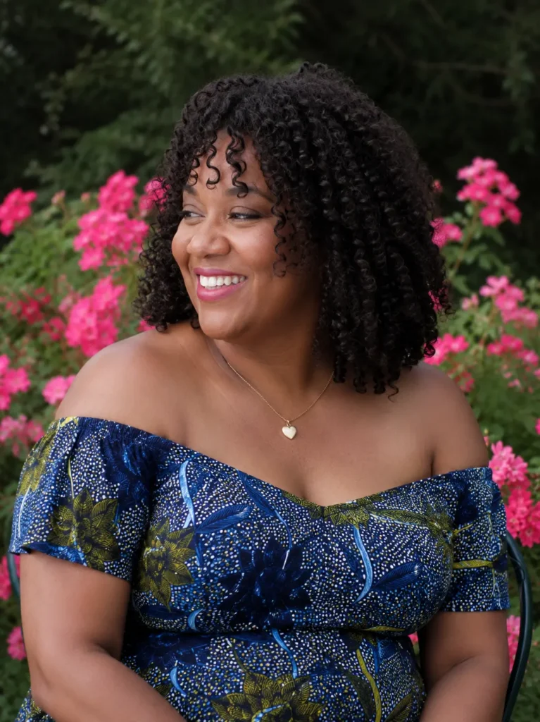Close-up portrait of a real plus-size woman in her 30s–50s with a defined spiral curly hairstyle featuring glossy, natural curls. She is wearing an elegant blue floral off-shoulder dress and smiling in an outdoor garden filled with vibrant pink flowers