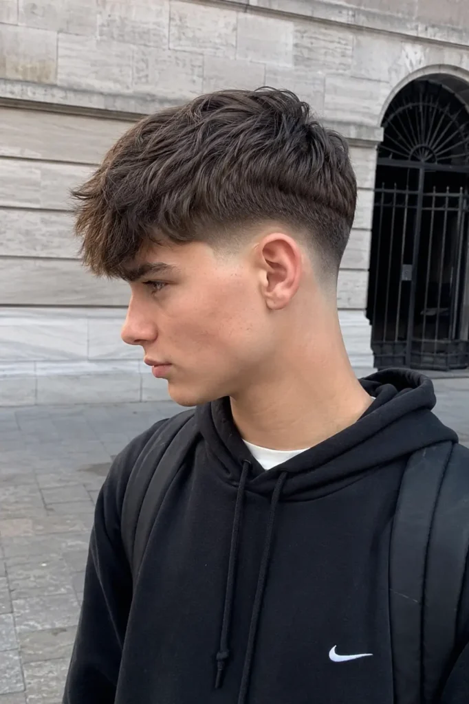 teenage boy with a modern Edgar haircut featuring a textured fringe and faded sides, wearing a black hoodie in an outdoor urban setting with natural daylight