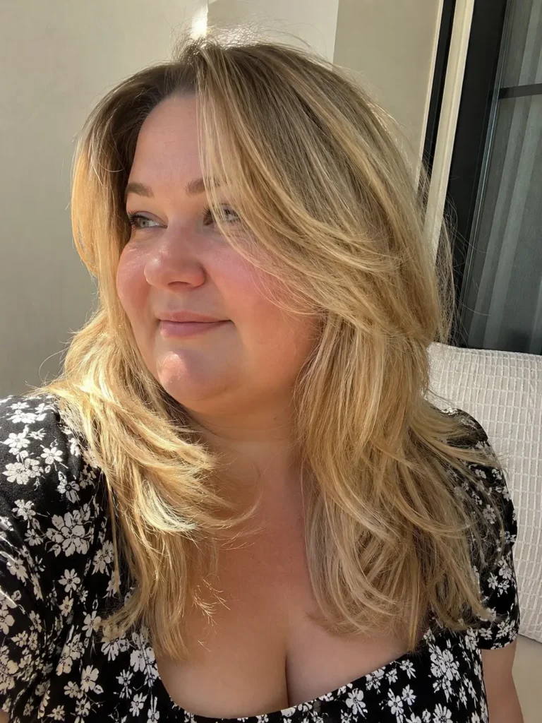 a plus-size English woman aged around 27–44 with a face-framing layered medium haircut in glossy golden blonde. She has natural skin texture and wears a black floral dress