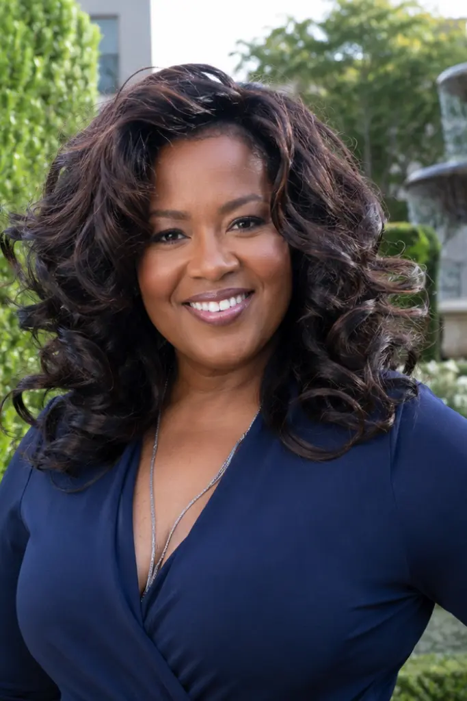 plus-size woman aged 30–60 with a glamorous curly blowout hairstyle featuring voluminous, shiny curls. She is smiling in an elegant navy dress in a luxury garden setting with manicured greenery and a fountain in the background