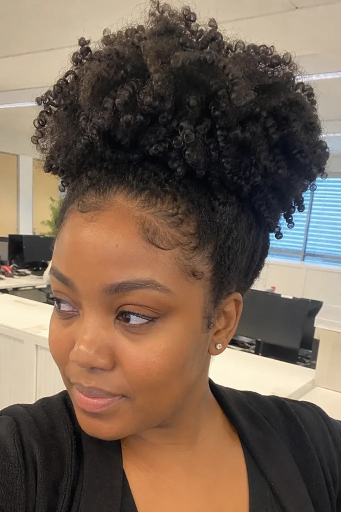 everyday woman with a natural high puff hairstyle featuring defined Type 4 curls and voluminous crown height, photographed in a modern office setting. The hairstyle showcases realistic curl definition, natural texture