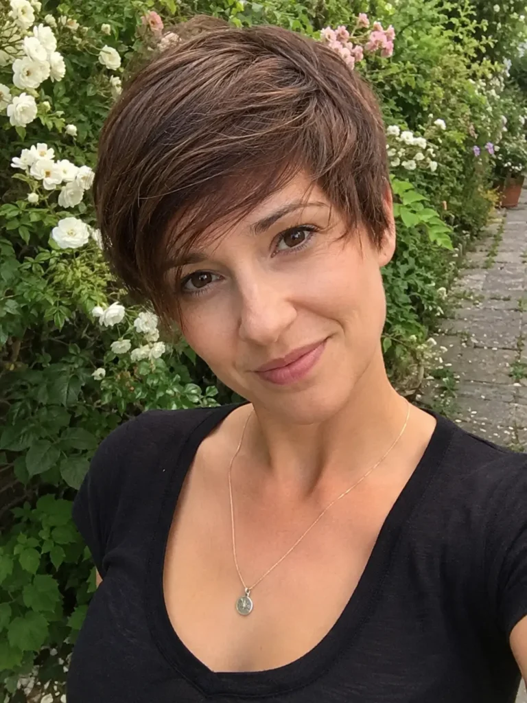 woman aged 25–40 with a layered pixie haircut and long side-swept bangs in chestnut brown hair, photographed outdoors in a blooming floral garden. She wears a casual chic black top and a delicate necklace