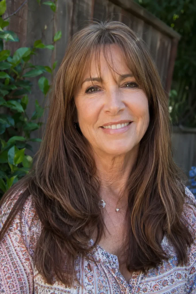 Mature woman aged 55–65 with long medium brown hair featuring wispy bangs and subtle highlights, smiling outdoors in natural daylight