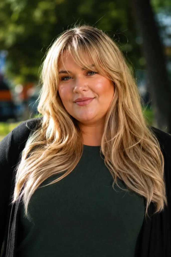 Plus-size English woman with long layered golden blonde hair and Bardot bangs, wearing a modest dark green top and black cardigan.