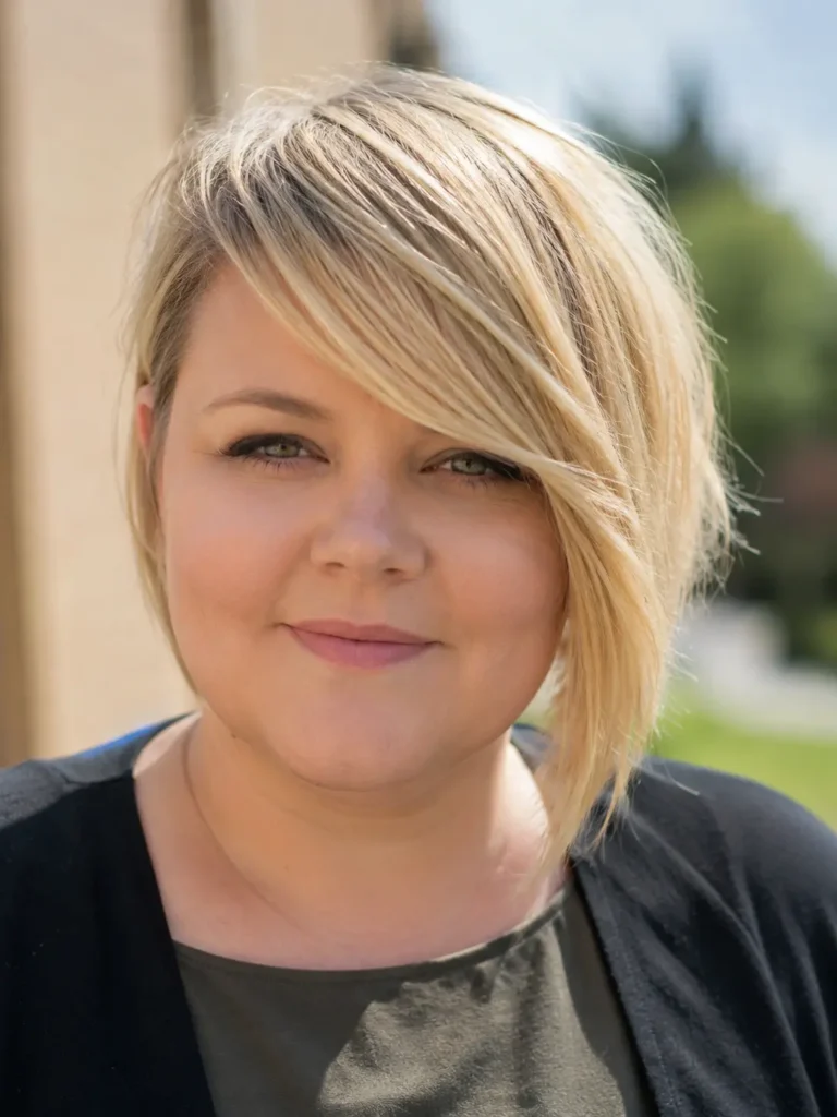 **Alt text:**
Real English woman with a round face wearing a long side-swept blonde pixie bob haircut, featuring a sweeping fringe, soft layers, and shiny hair, photographed in natural sunlight with a realistic everyday appearance.
