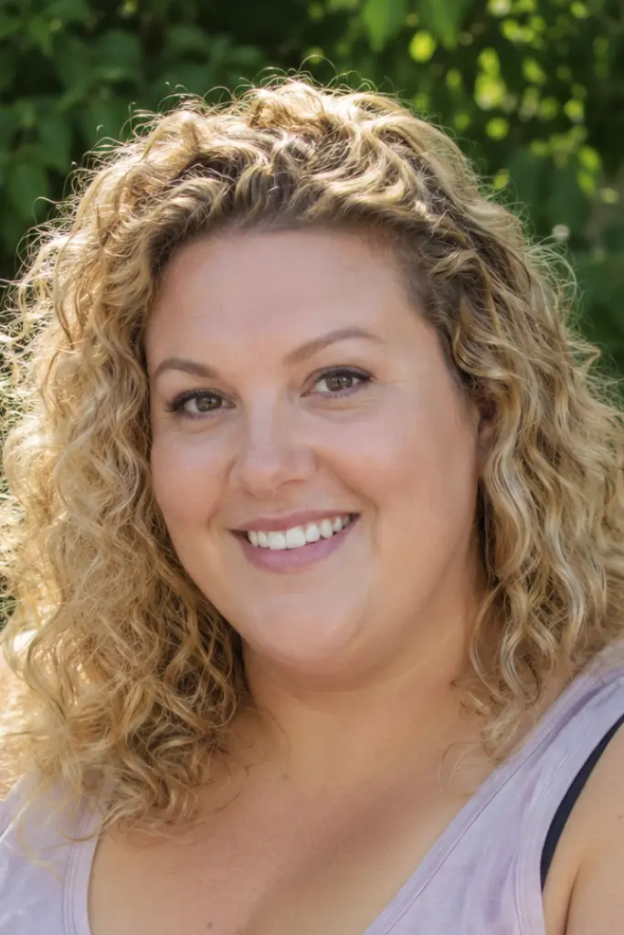 English plus-size woman with medium-length loose curly beige blonde hair featuring voluminous crown lift and defined curls, smiling outdoors in bright natural daylight