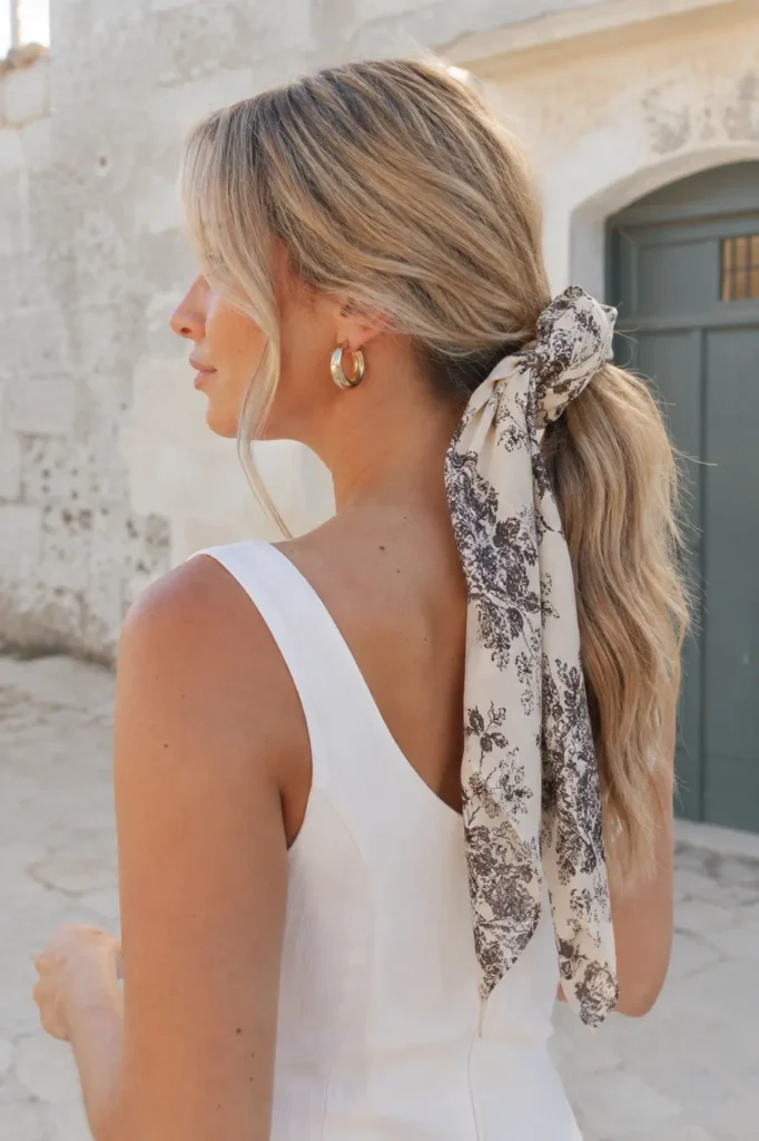 
English woman with honey blonde low ponytail tied with a patterned bandana bow in natural outdoor light.
