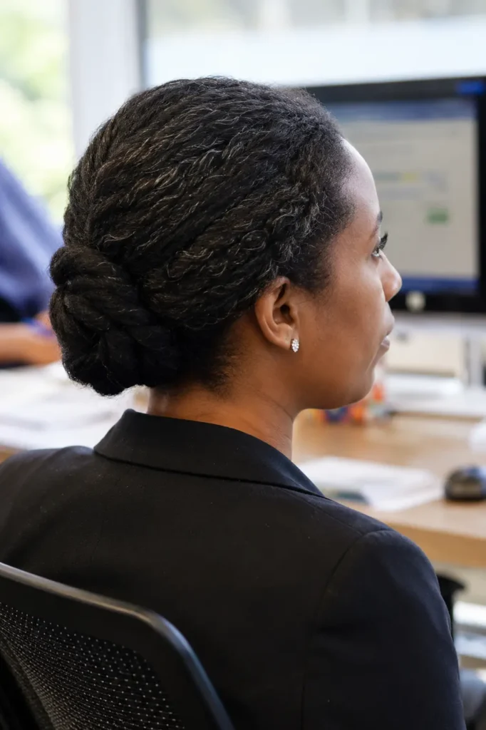 Back and side profile of a real woman in a corporate office wearing a low twisted bun hairstyle on natural hair. The hairstyle is shown in sharp focus, highlighting the twisted bun detail, natural texture, and smooth styling. She wears professional business attire and sits in a bright office setting with soft daylight
