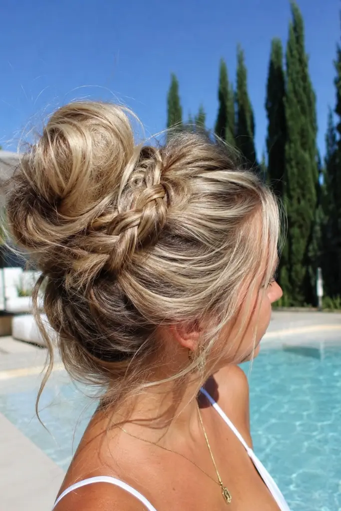 woman aged 25–45 with light golden blonde hair styled in a messy braided bun, photographed from a side-back angle beside a luxury swimming pool. The intricate braid wraps into a textured, relaxed bun with loose wisps framing the hairstyle