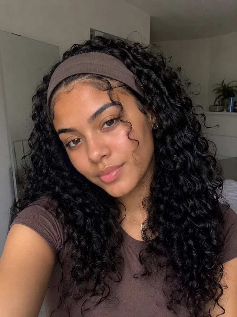 Realistic portrait of a 21-year-old woman wearing a neutral fabric headband with loose natural curls cascading around her face. Her authentic facial features and natural skin texture are highlighted in soft, realistic lighting