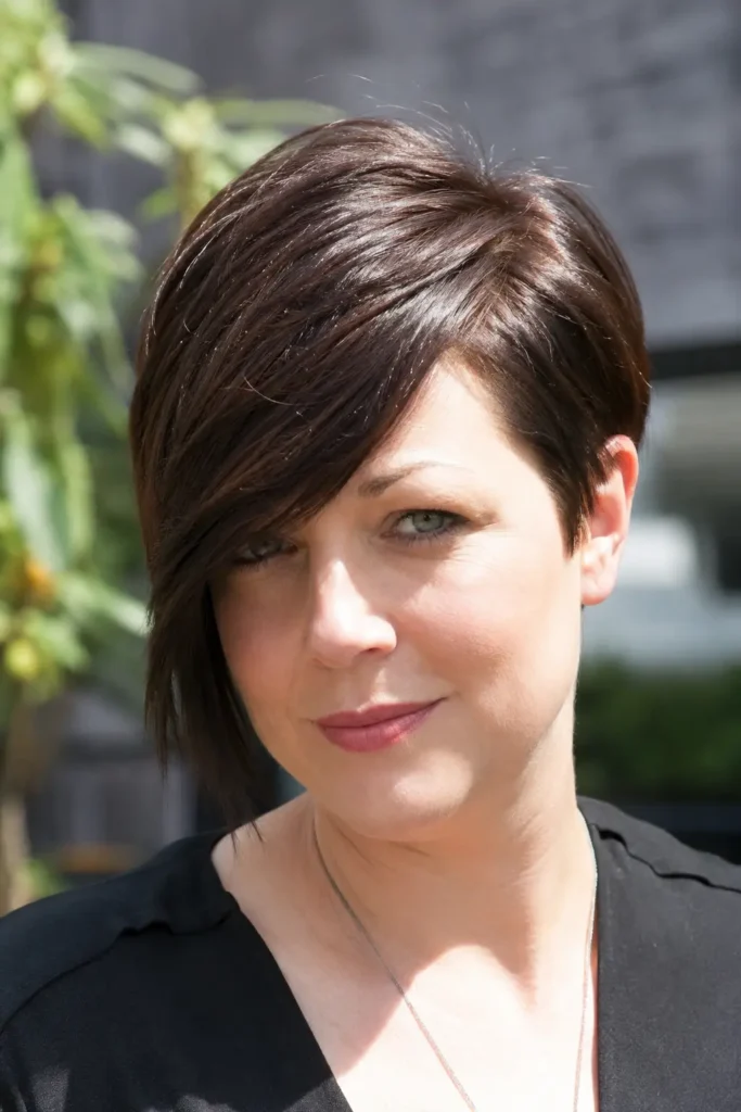 woman with a round face and a pixie bob haircut featuring a long tapered side fringe. The glossy dark brown hair is styled with a sweeping fringe that frames the face, while the tapered back and sides create a modern silhouette
