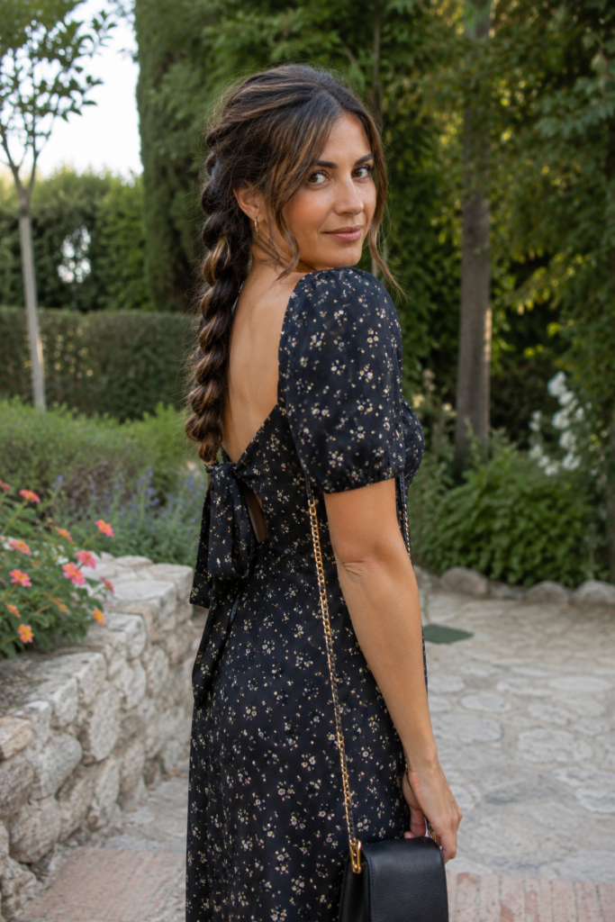 Back view of a real woman aged 25–40 with a pull-through braid and long brunette hair with subtle highlights, wearing a black floral dress in a lush garden setting