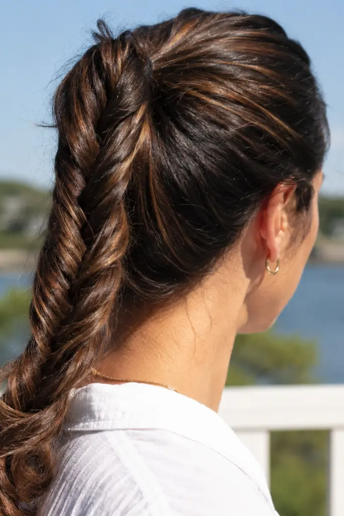 A woman with a brunette rope twist ponytail stands outdoors in bright summer sunlight, viewed from the side and slightly behind