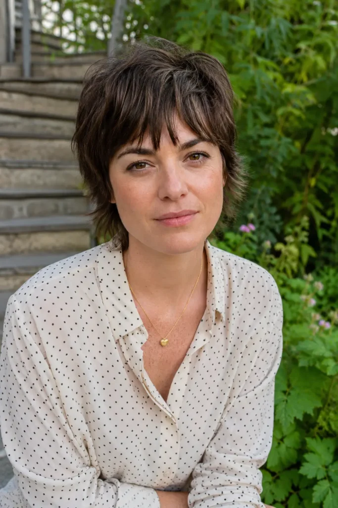 woman with a shaggy pixie haircut and feathered bangs, featuring medium brown textured hair and natural skin texture, wearing a casual chic blouse in an outdoor garden setting