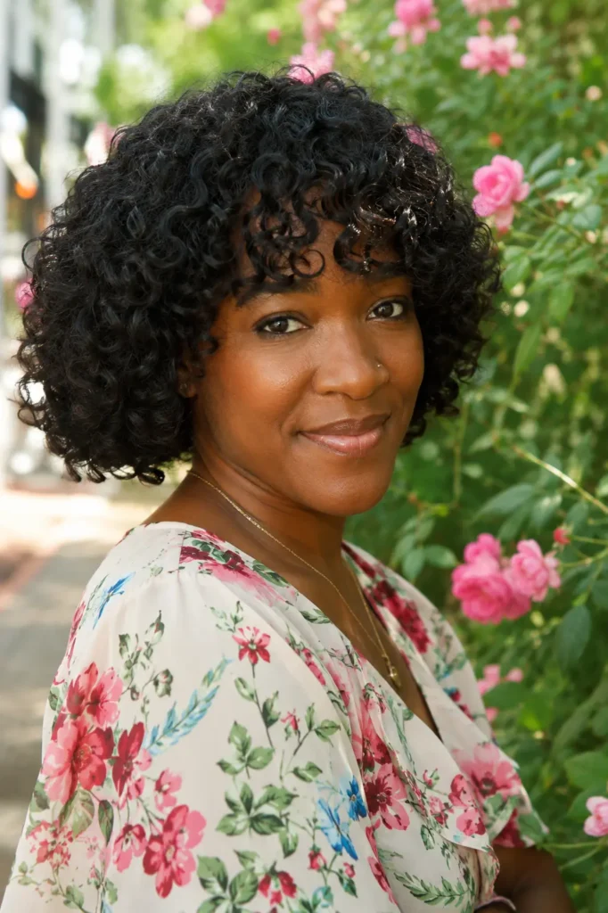 Black woman with a short curly bob and curly bangs, featuring defined natural curls and realistic skin texture, wearing a floral blouse in an outdoor garden filled with pink flowers