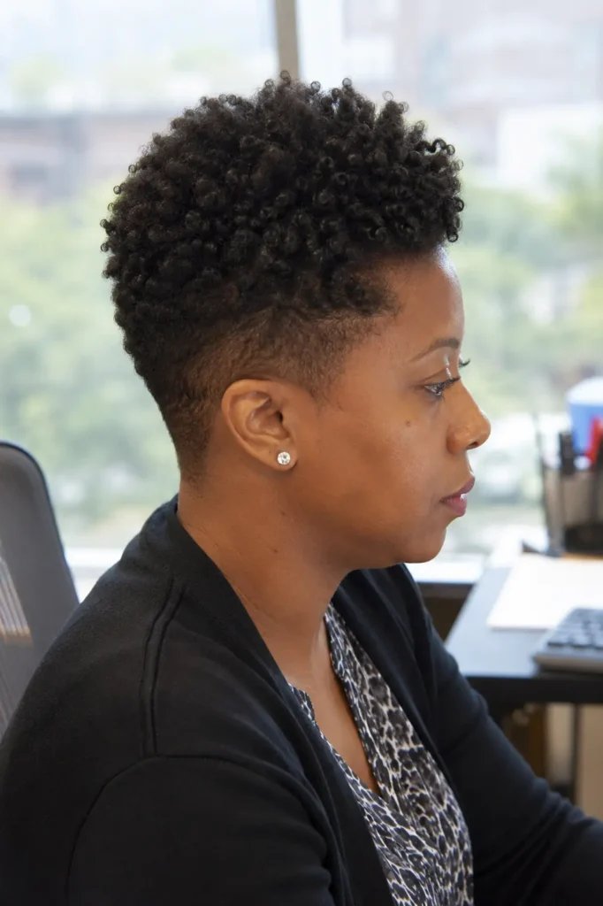 woman with a short tapered natural haircut featuring defined Type 4 curls on top, shown in side profile while working in a professional office setting. The hairstyle is prominently displayed, highlighting the tapered sides, natural curl texture, and realistic hair density