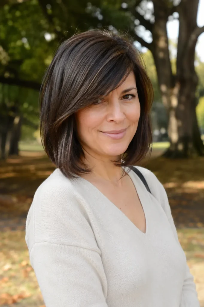 woman aged 30–50 with a shoulder-length bob haircut and side-swept bangs, showcasing glossy dark brunette hair. She wears an elegant casual outfit and poses outdoors in a lush park setting