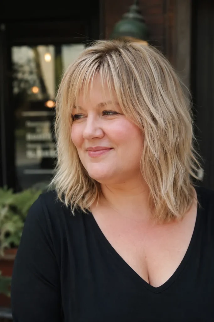 Plus-size English woman with a shoulder-length shag haircut and wispy bangs, featuring light beige blonde hair with textured layers and healthy shine