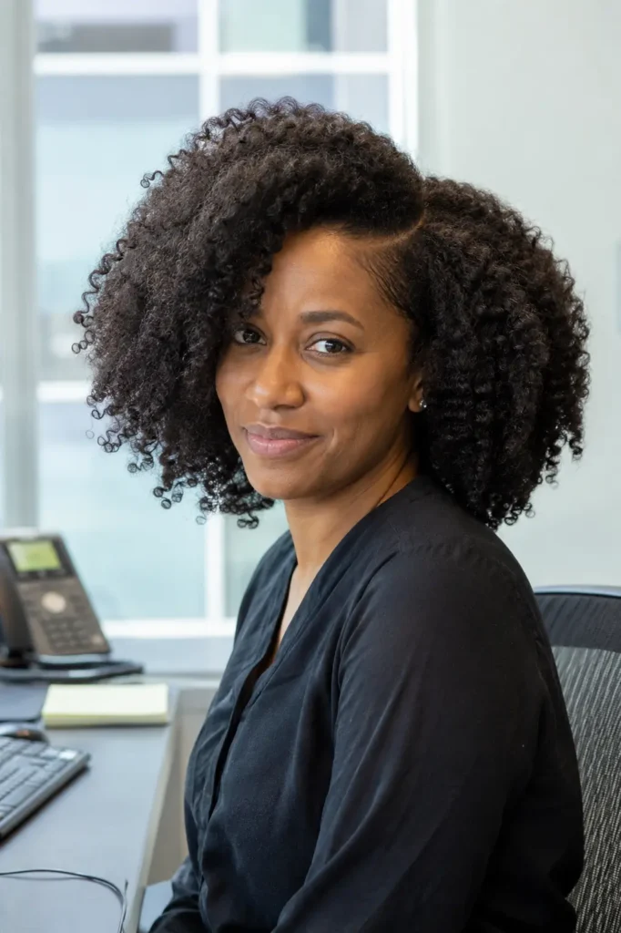 woman in a professional office setting wearing a black blouse, featuring a voluminous side-part curly afro hairstyle with defined natural curls and realistic texture. Soft daylight highlights the shape, density, and volume of the hair