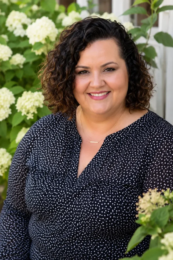 a real plus-size woman with a side-part curly bob hairstyle, featuring defined, voluminous curls framing her face. She wears a stylish dark blouse and smiles warmly in an outdoor floral garden setting