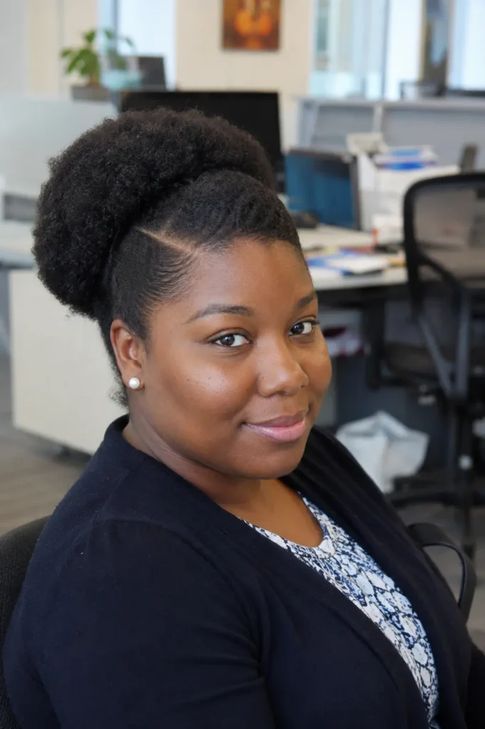 everyday Black woman wearing a side-part puff hairstyle with natural Type 4 hair, featuring a defined side part, sleek edges, and a voluminous curly puff. She is seated in a professional office environment with a softly blurred workplace background