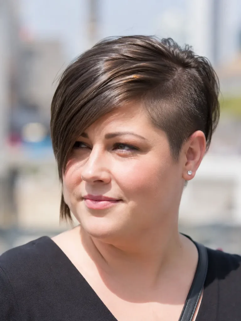 woman with a round face wearing a side-shaved pixie bob haircut featuring asymmetrical styling and a long side-swept fringe.