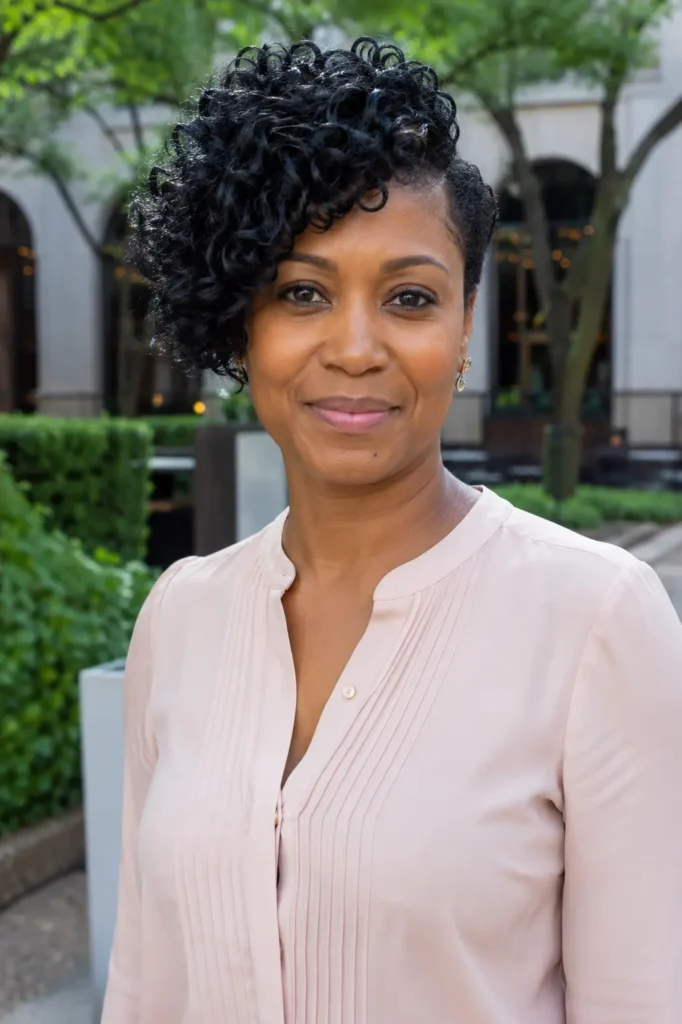 Black woman with a side-swept curly crop hairstyle featuring defined natural curls and a tapered side. She wears an elegant light blouse and stands outdoors in a city garden setting with softly blurred greenery and urban architecture in the background.