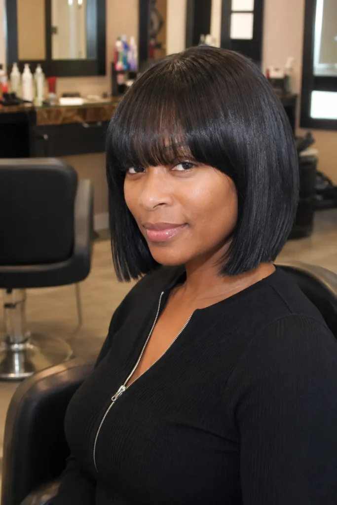 woman aged 25–45 with a sleek silk press bob haircut and full bangs, featuring glossy natural black hair. She is seated in an indoor salon setting, wearing an elegant black outfit