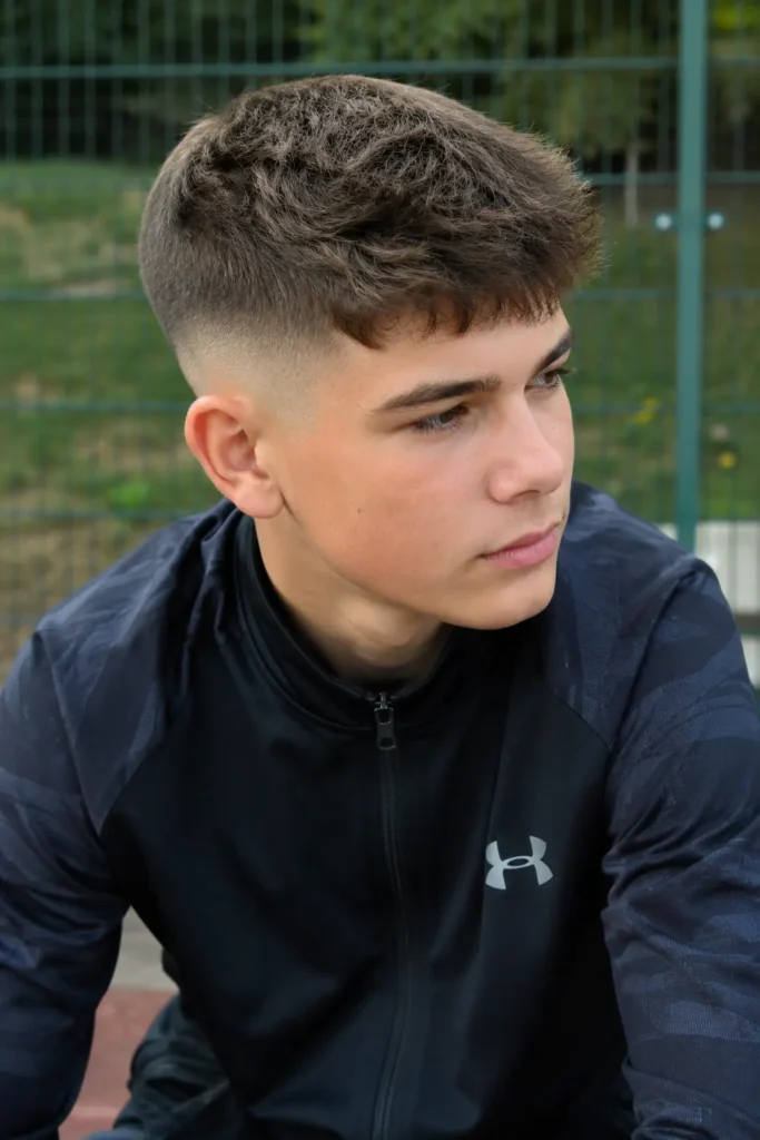 teenage boy with a skin fade haircut and short textured top, wearing dark sportswear in an outdoor setting. The sharp fade blends smoothly to the skin, while natural daylight highlights his realistic facial features, healthy hair texture, and authentic teenage appearance