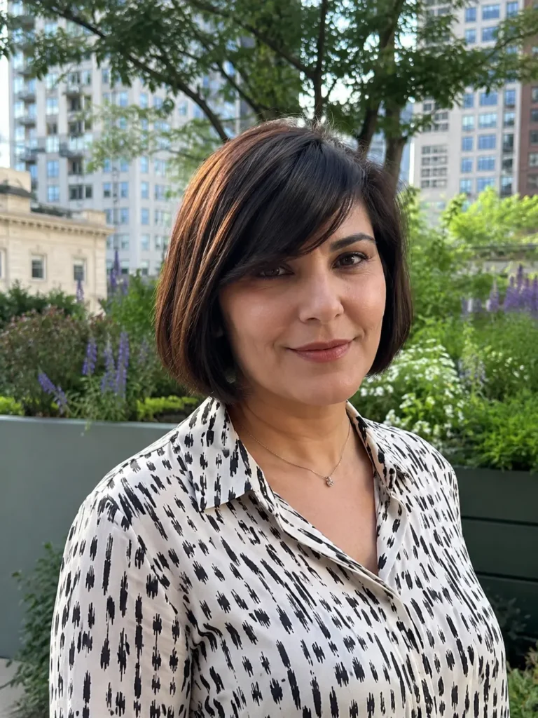 woman aged 28–48 with a sleek A-line bob haircut and side-swept bangs, featuring glossy dark brown hair. She wears an elegant patterned blouse and stands in an outdoor city garden with lush greenery and softly blurred urban buildings in the background